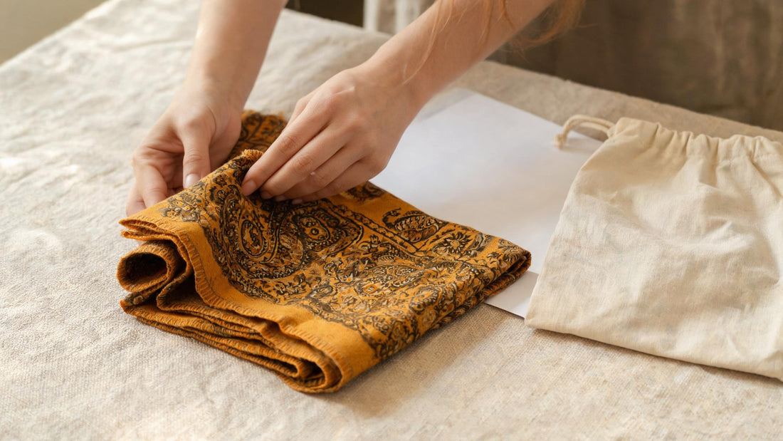 Hands folding a saffron paisley shawl on a linen-covered table beside tissue paper and a cotton storage bag