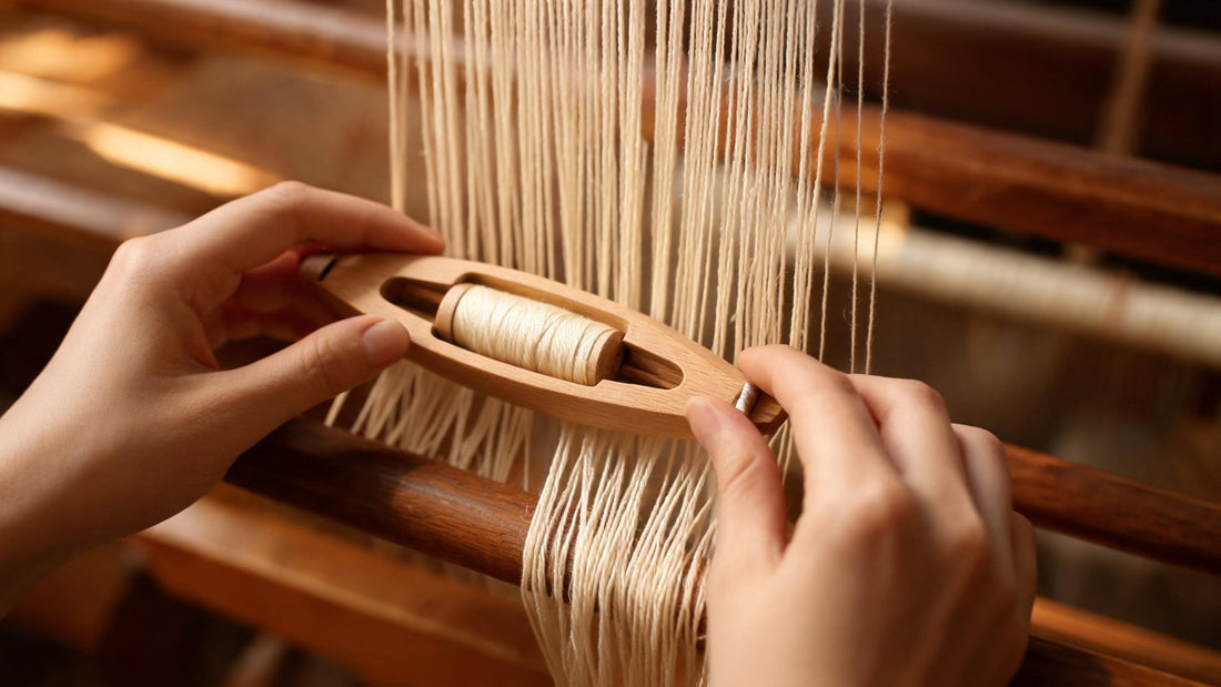 Weaver's hands passing a wooden shuttle through fine cream warp threads on a handloom