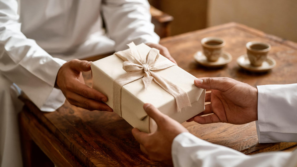 Two men in white thobes passing a gift box tied with linen ribbon across a majlis table