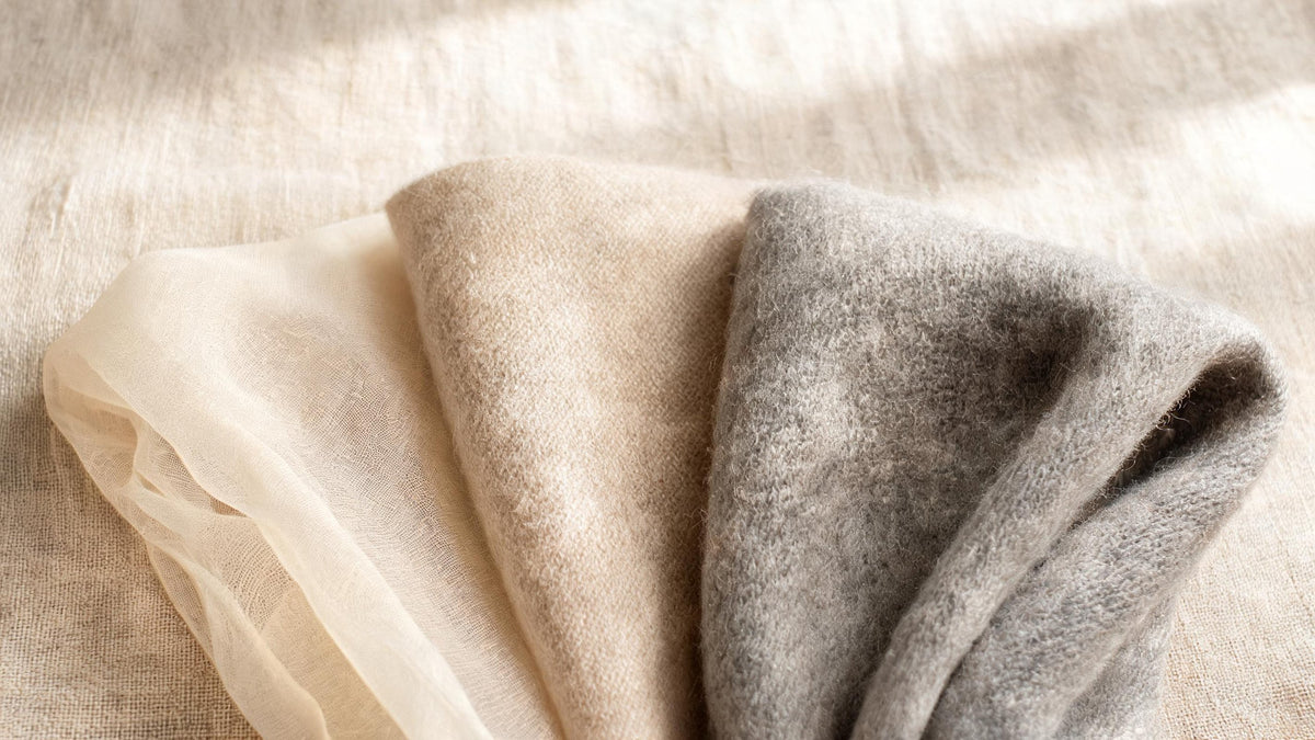 Three fabric swatches fanned out on linen: a sheer cream pashmina, an oatmeal blend and a heavier grey cashmere